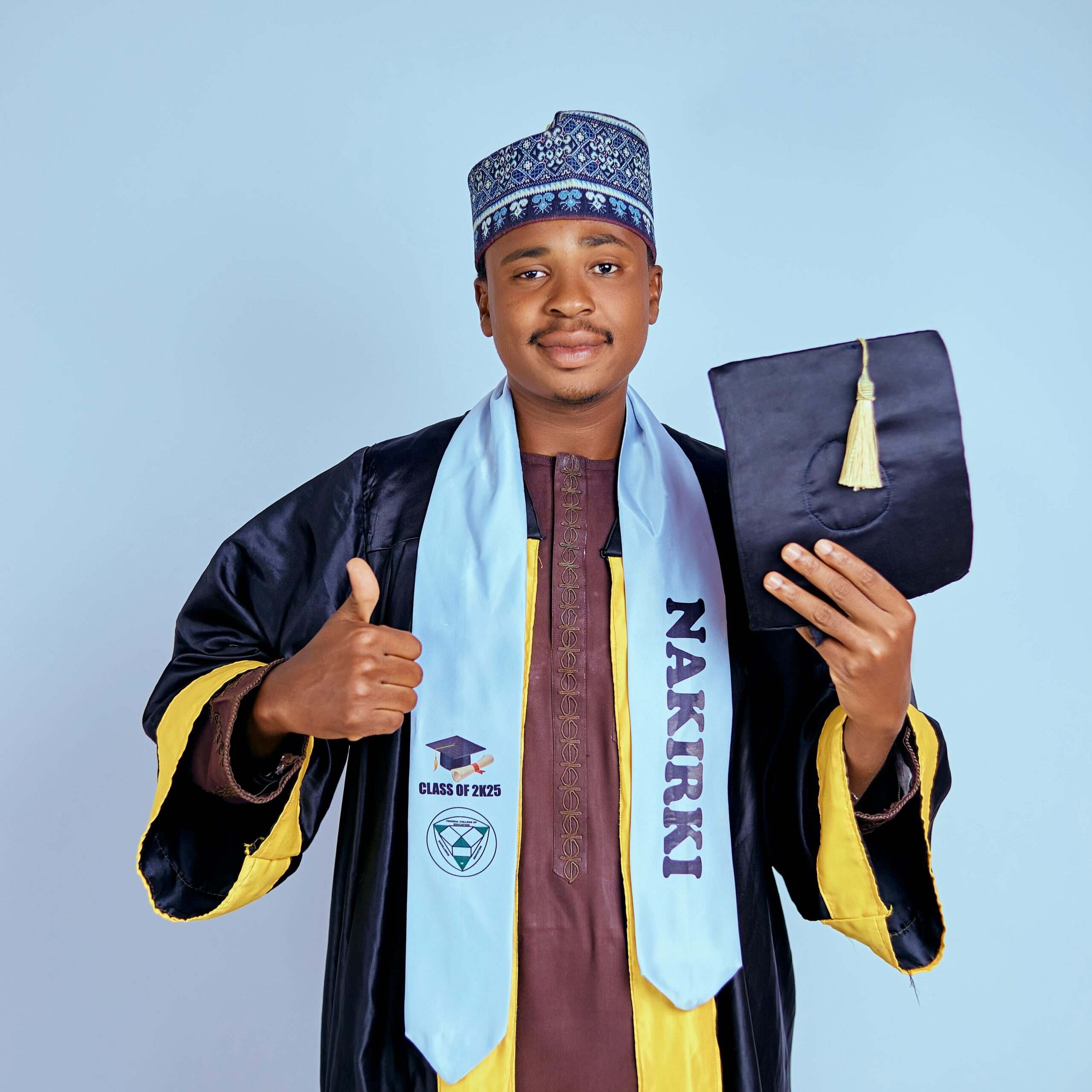 Portrait of a young graduate in traditional attire holding diploma and giving thumbs up.